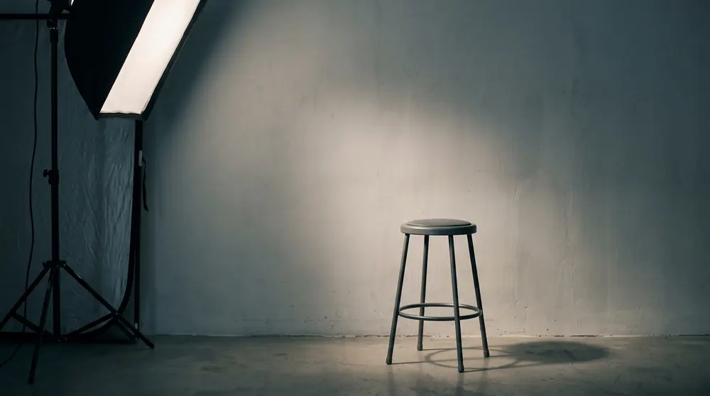 A single softbox raking soft light across a plain neutral wall with an empty presenter stool standing in the pool of light