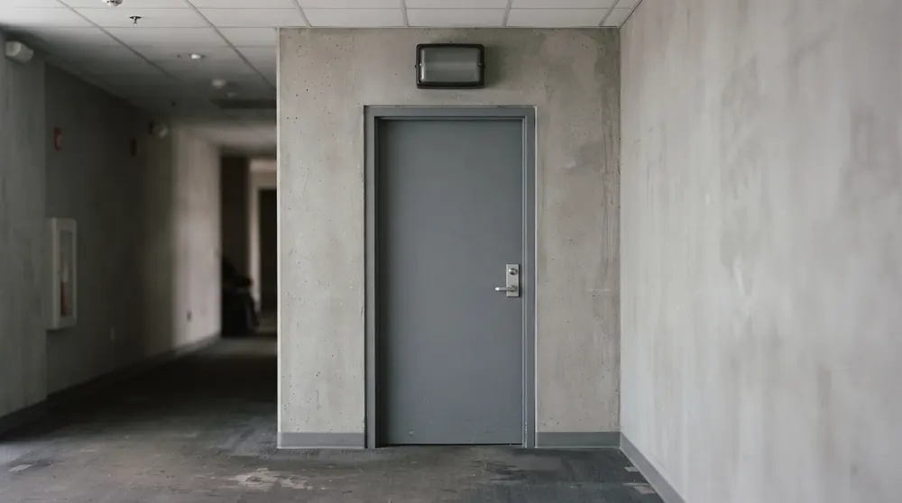 A plain closed studio door with a simple handle and an unlit signal lamp above it in a neutral corridor
